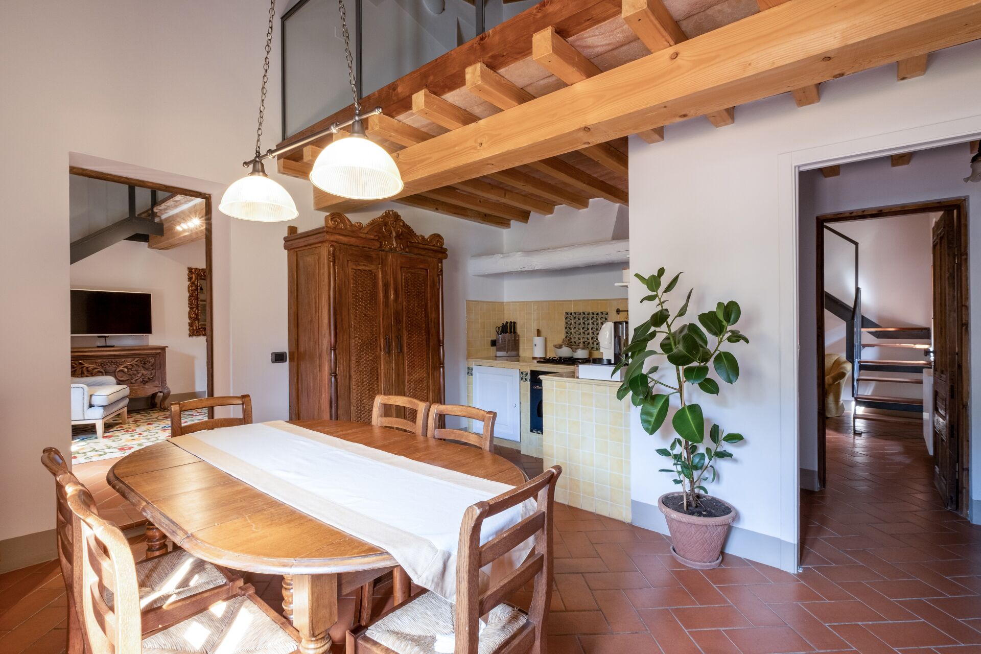 A dining area with a wooden table and chairs, a potted plant, and a kitchen in the background.