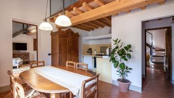 A dining area with a wooden table and chairs, a potted plant, and a kitchen in the background.
