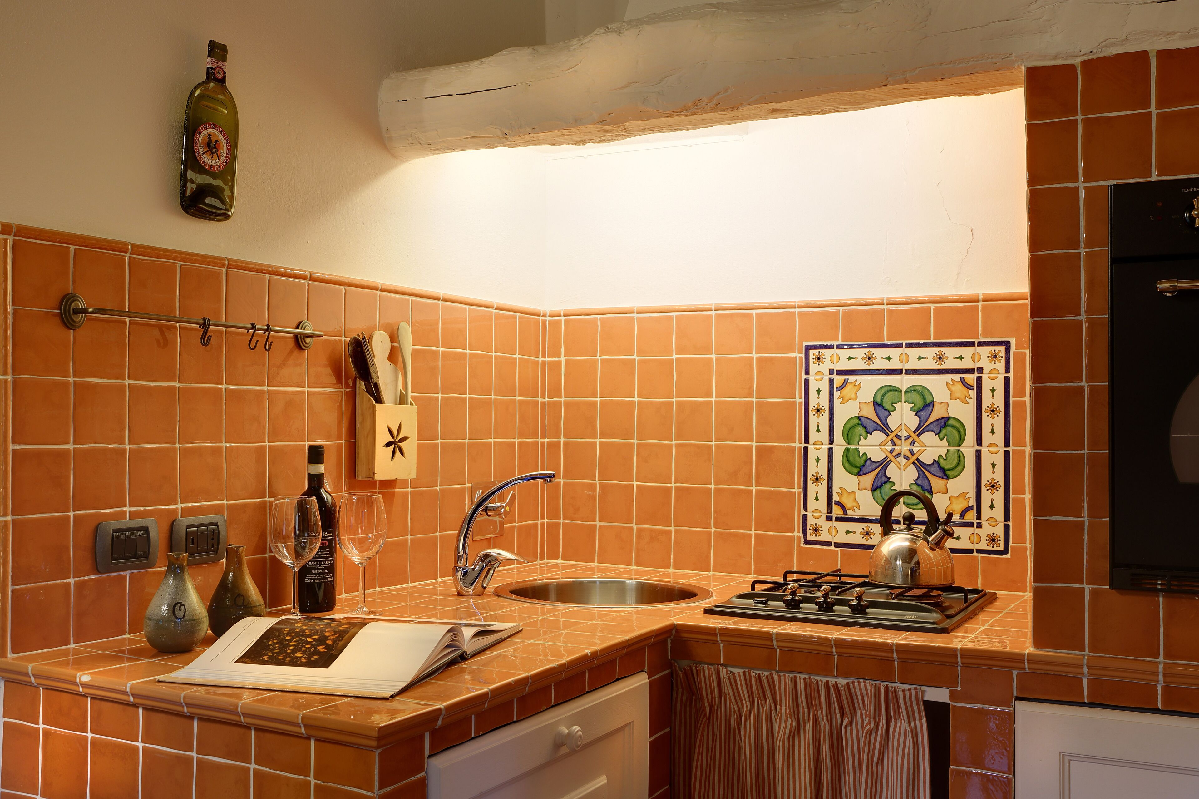Une cuisine avec des carreaux orange, un évier en acier inoxydable, une cuisinière à gaz et un mur recouvert de carreaux décoratifs.