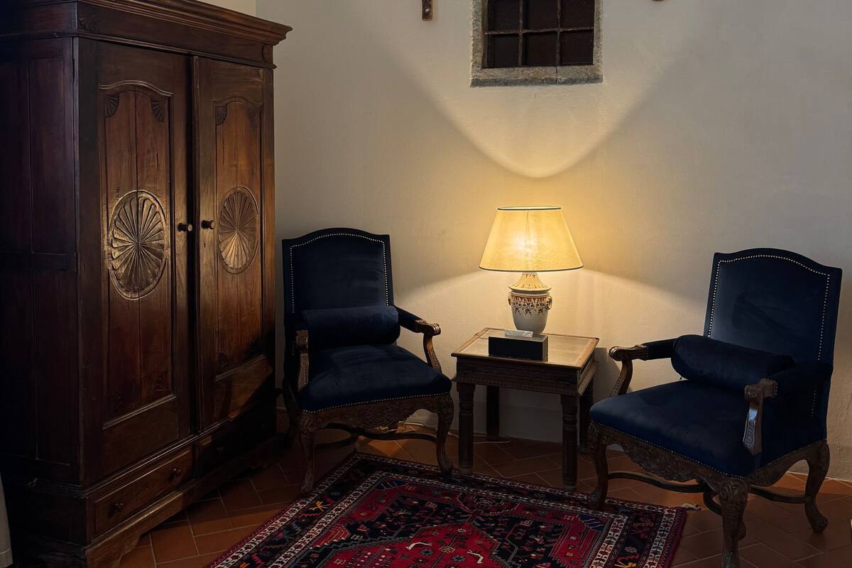 A room with a dark wooden cabinet, two blue upholstered chairs, a small table with a lamp, a decorative wall piece, and a red patterned rug.