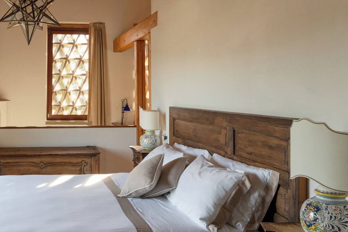A bedroom with a wooden ceiling, a bed with white linens, a nightstand with a lamp, and a window with curtains.