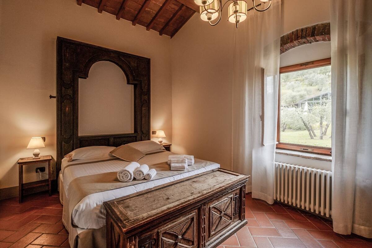 A bedroom with a large bed, two bedside lamps, a wooden nightstand, a radiator, and a window with curtains.