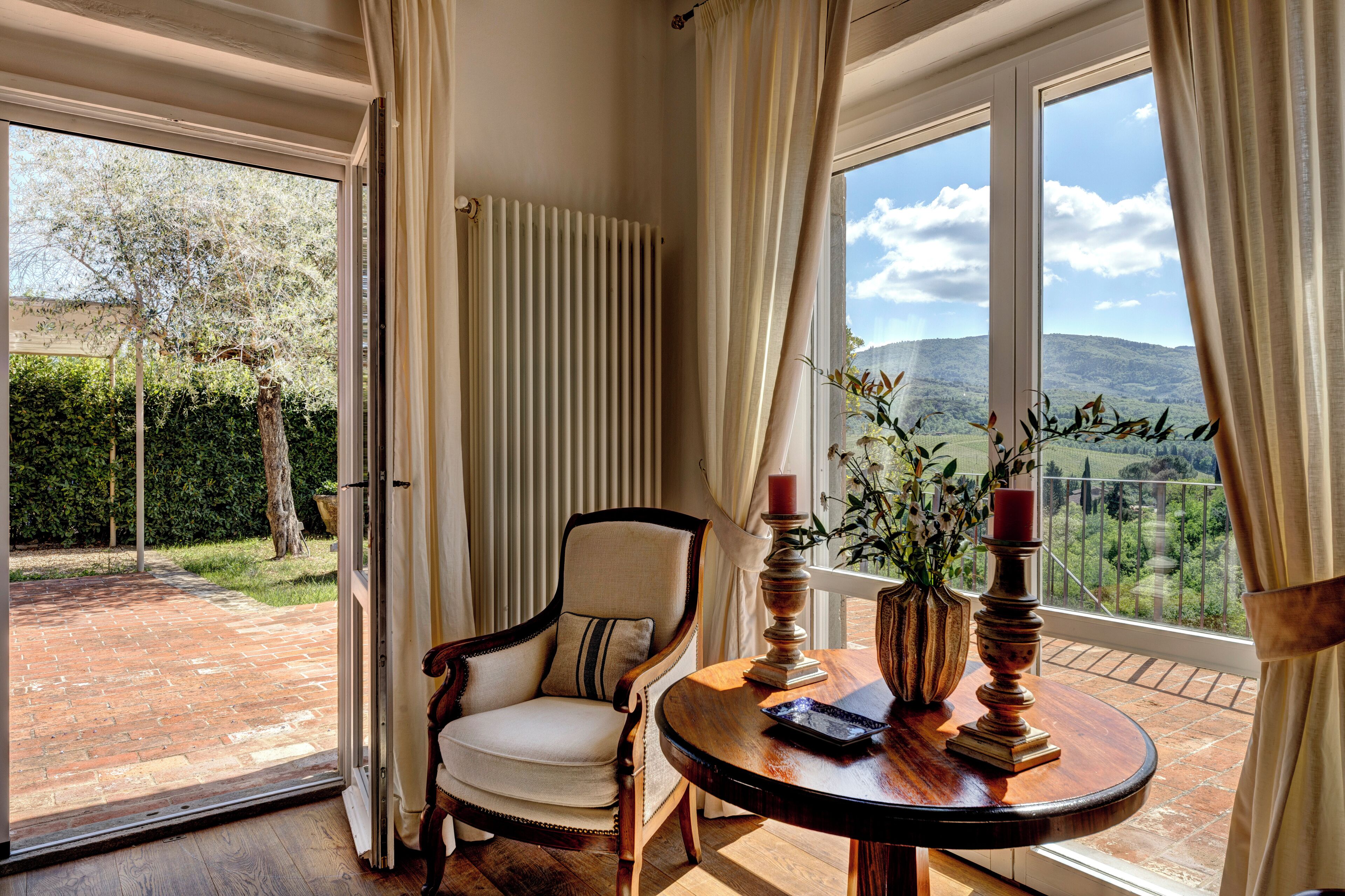Quarto com vista para uma paisagem verde, uma cadeira, uma mesa com uma vela e um vaso com flores.