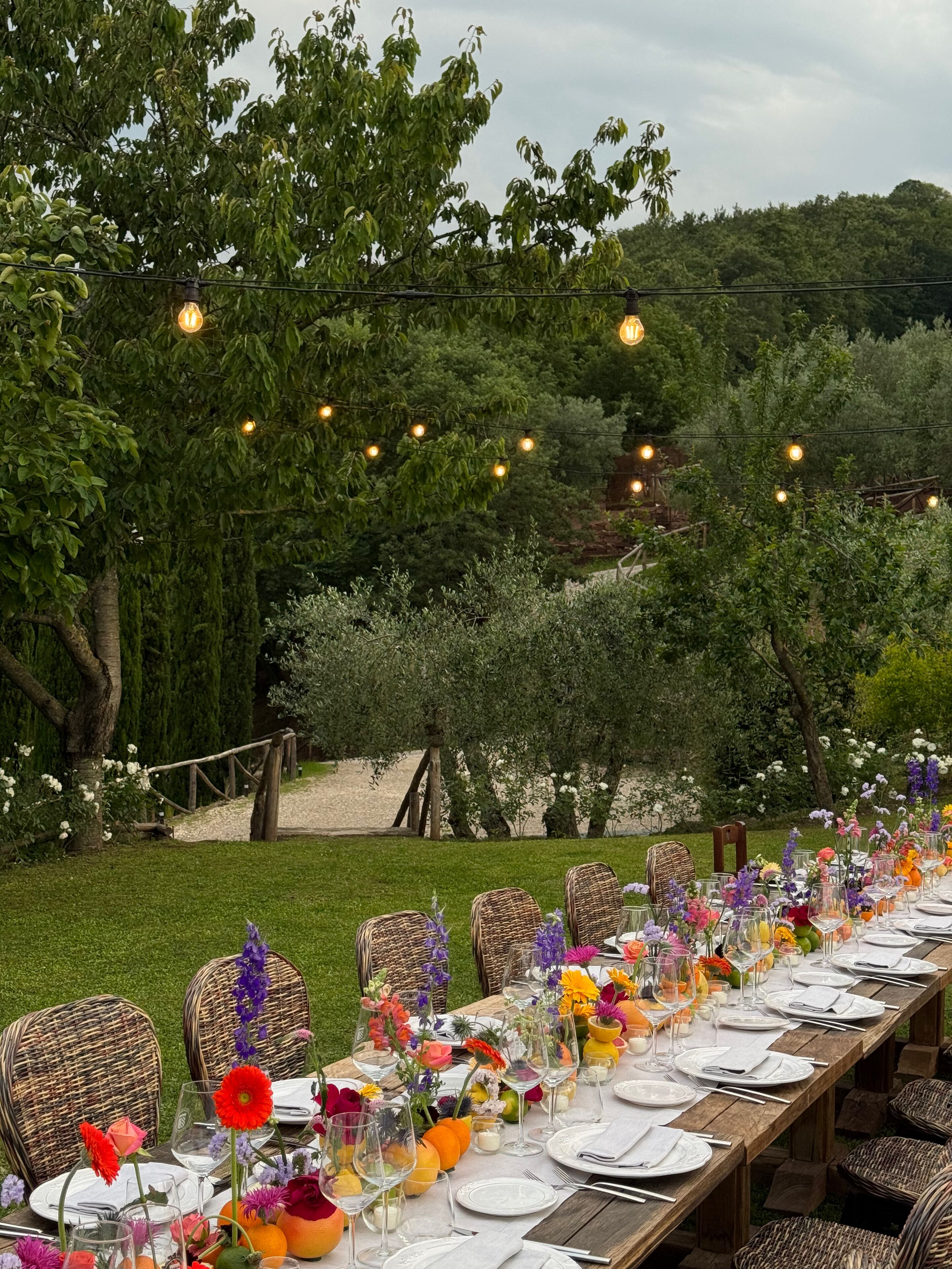 An outdoor dining area with a long wooden table set for a meal, surrounded by wicker chairs, under string lights.