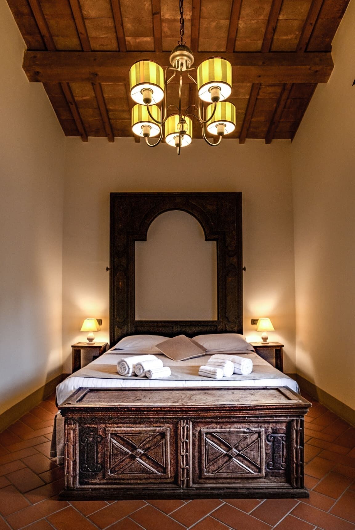 A bedroom with a wooden bed frame, a large mirror above the bed, and a chandelier with four lamps.