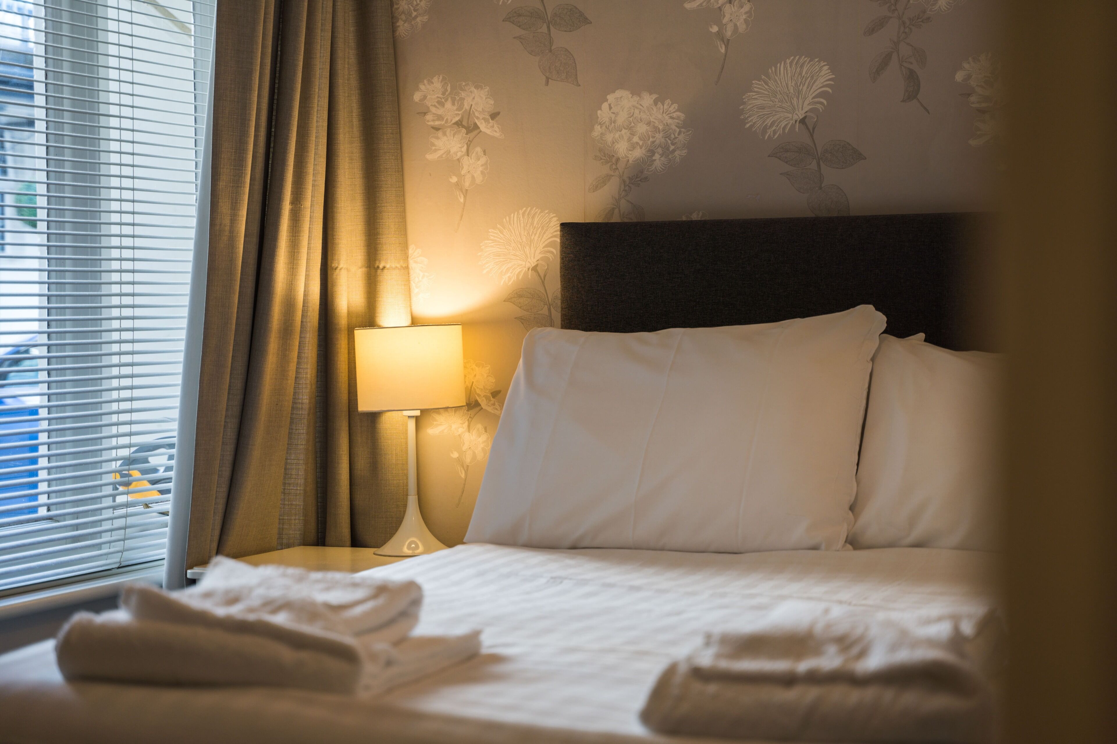 A neatly made bed with white linens, a bedside lamp, and a window with blinds.