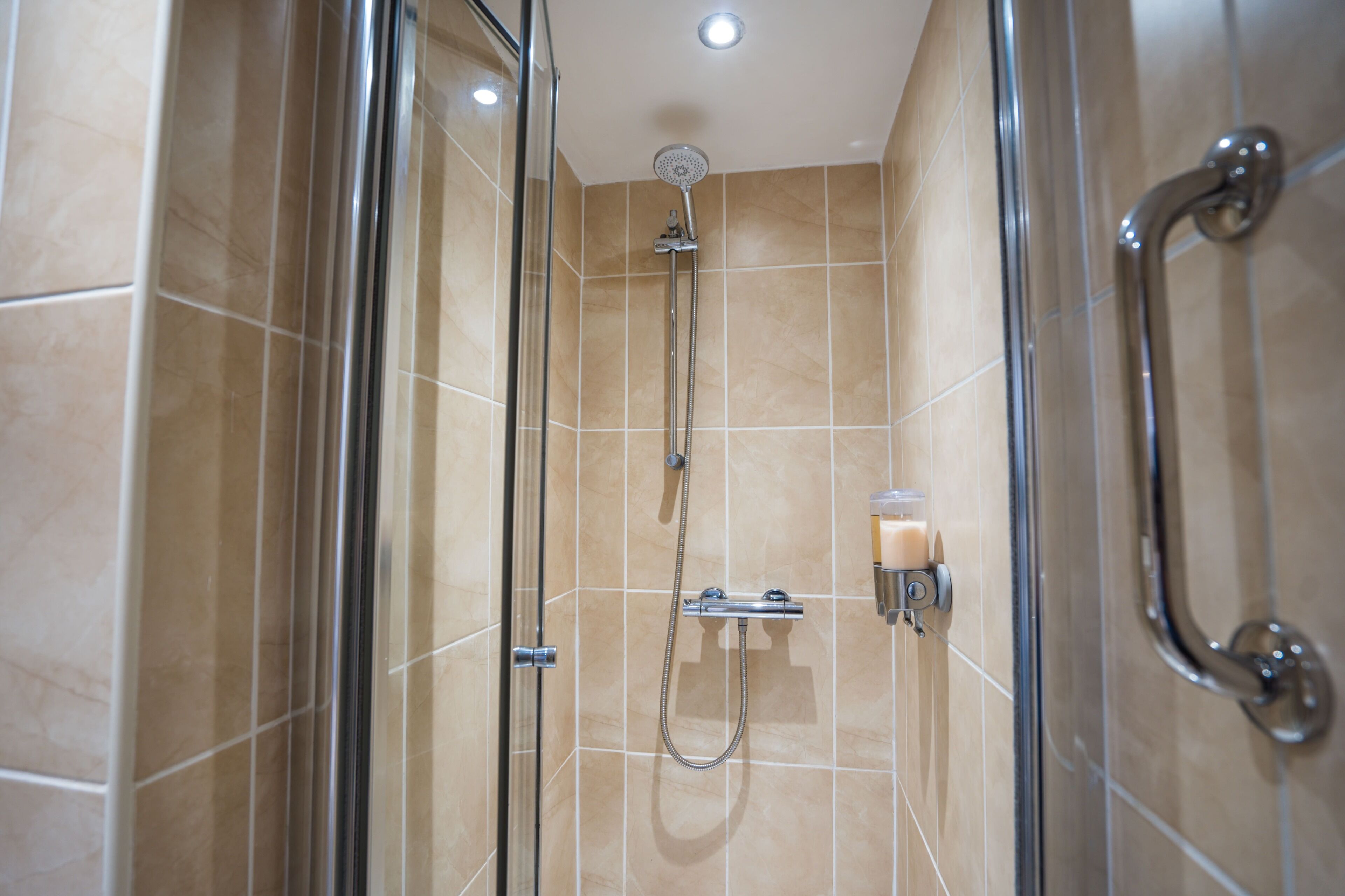 Ducha con cerramiento de vidrio, cabezal de ducha manual y dispensador de jabón en la pared.