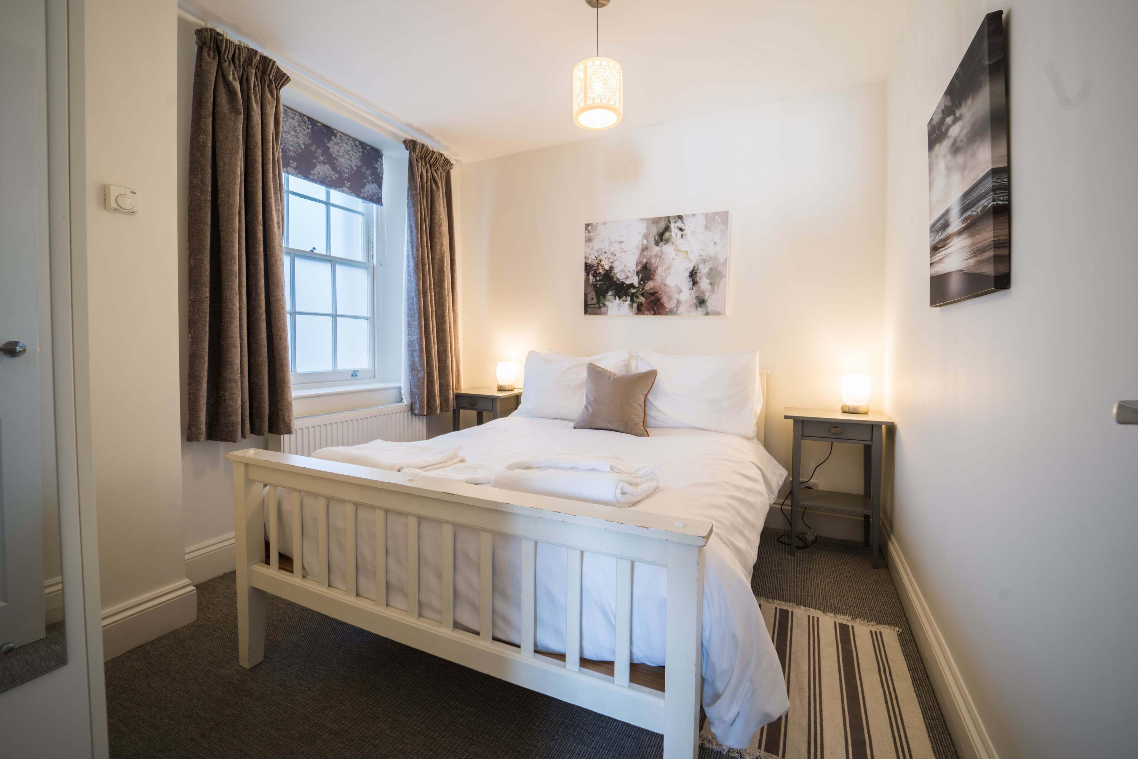A bedroom with a white bed, a window with curtains, a bedside table with a lamp, and a wall-mounted picture.