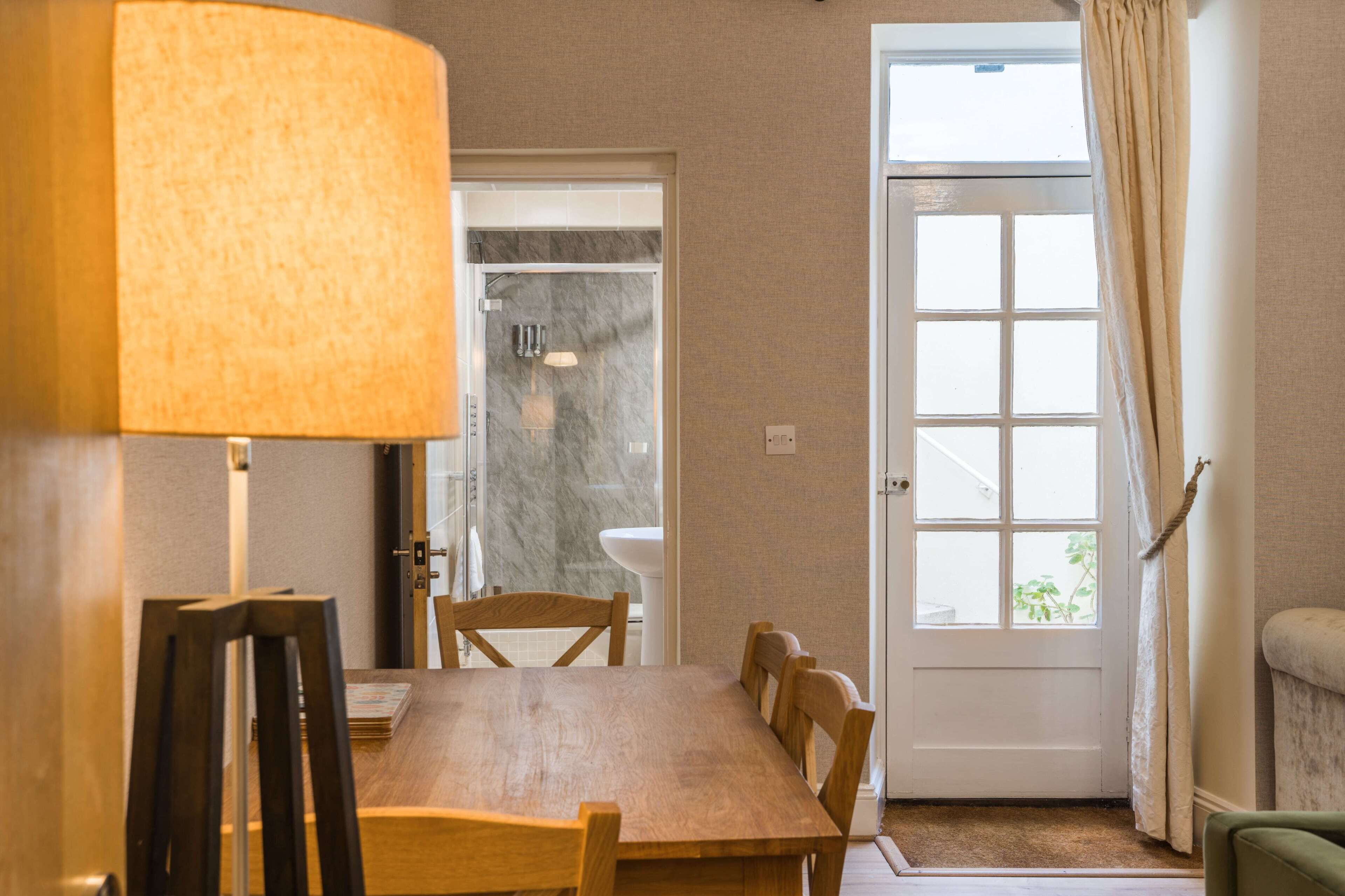 A dining area with a wooden table and chairs, a lamp, a door leading to a bathroom, and a window with curtains.