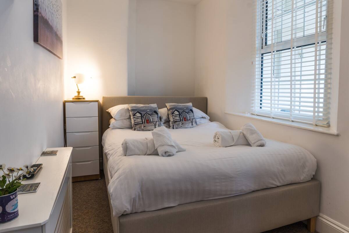 A neatly made bed with pillows and towels, a bedside table with a lamp, a desk with a plant, and a window with blinds.