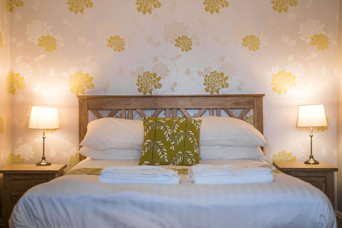A neatly made bed with white linens and a green leaf-patterned pillow, flanked by two bedside tables with lamps, set against a floral wallpaper background.