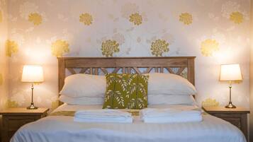 Una cama bien hecha con ropa blanca y un cojín estampado con hojas verdes, flanqueada por dos mesitas de noche con lámparas, sobre un fondo de papel tapiz floral.