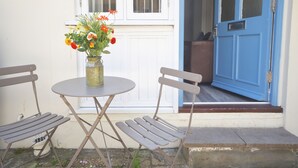 Een klein buitenzitje met een tafel en twee stoelen, een vaas met bloemen en een blauwe deur die naar een kamer leidt.