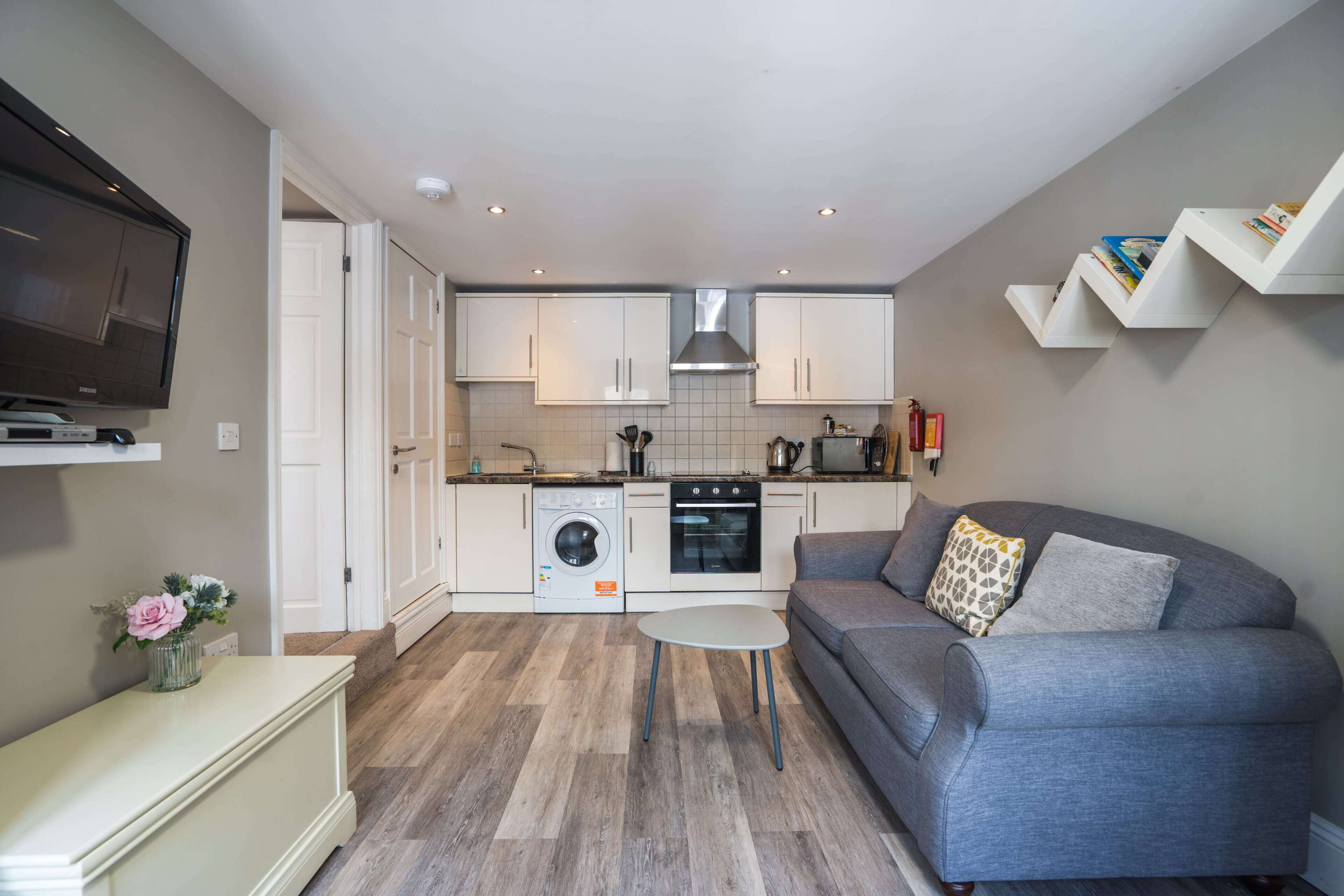 Un espacio habitable compacto con kitchenette, un sofá gris, una mesita y un televisor montado en la pared.