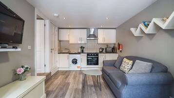 Un espacio habitable compacto con kitchenette, un sofá gris, una mesita y un televisor montado en la pared.