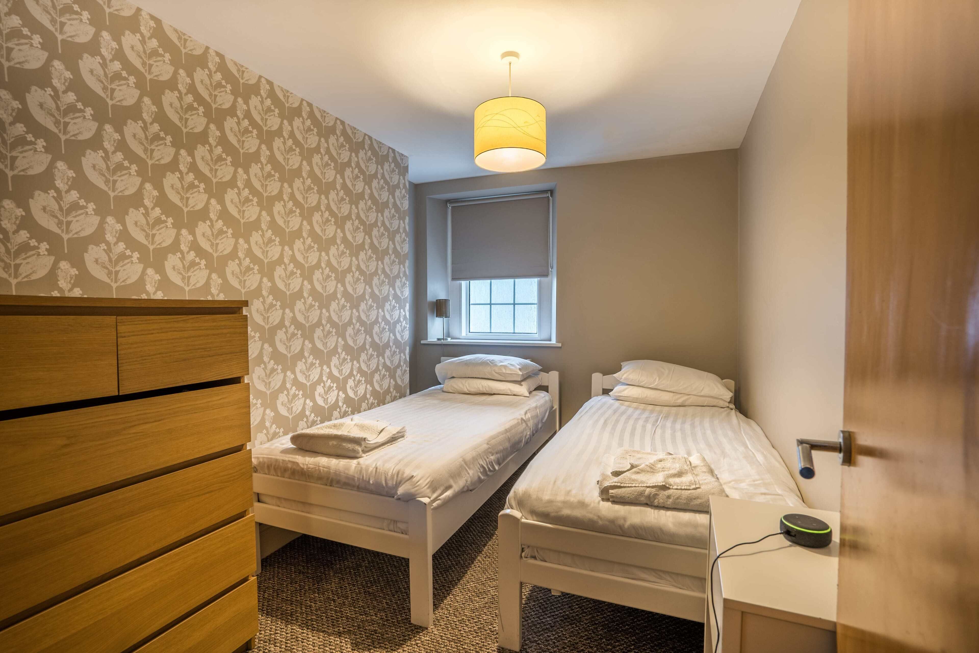 Un dormitorio con dos camas individuales, un aparador de madera, una ventana con persiana y una lámpara en el techo.