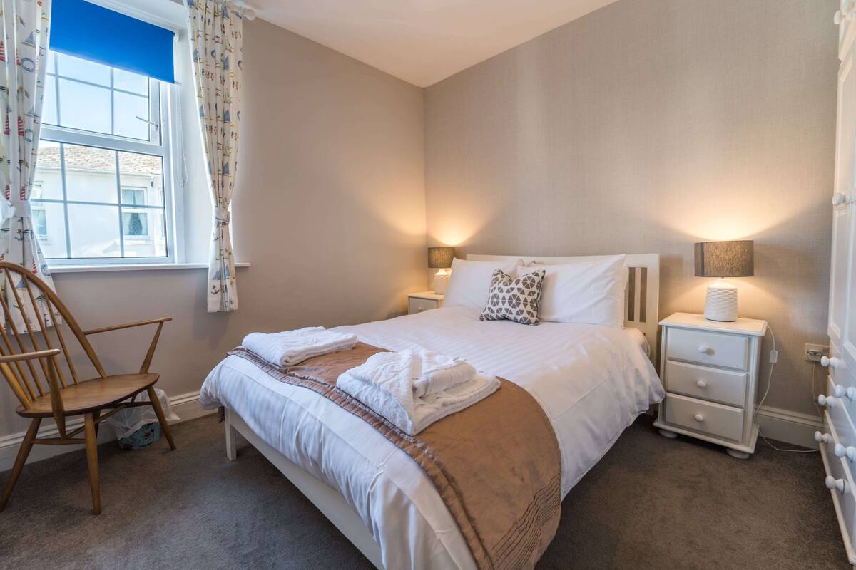 A neatly made bed with white linens and a brown throw blanket, a wooden chair, two bedside tables with lamps, and a window with curtains.