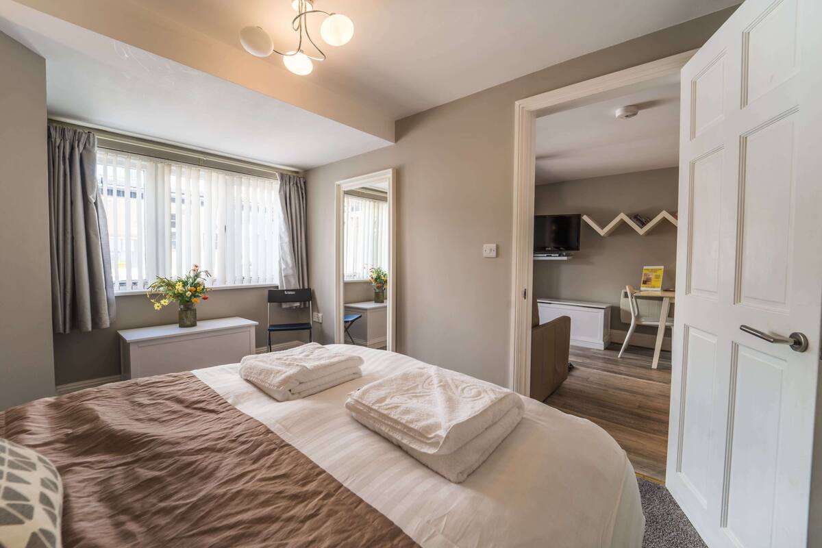 A neatly arranged bedroom with a large bed, a desk, and a television in the adjacent room.