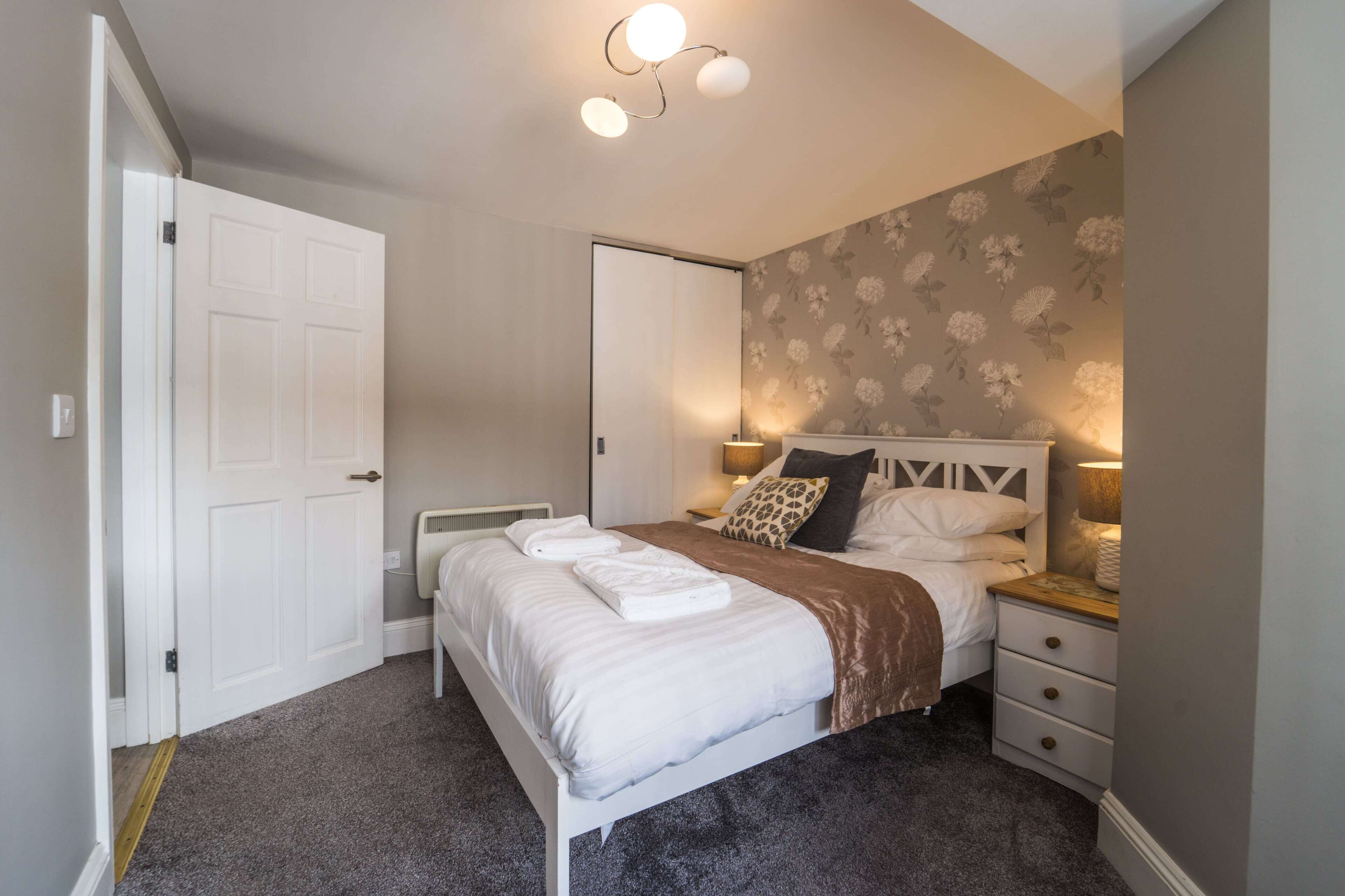 Une chambre à coucher comprenant un lit, des tables de chevet, une commode, une lampe fixée au mur et une porte.