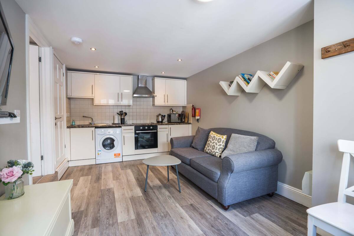 A compact living space with a kitchenette, a grey sofa, a small round table, and a washing machine.