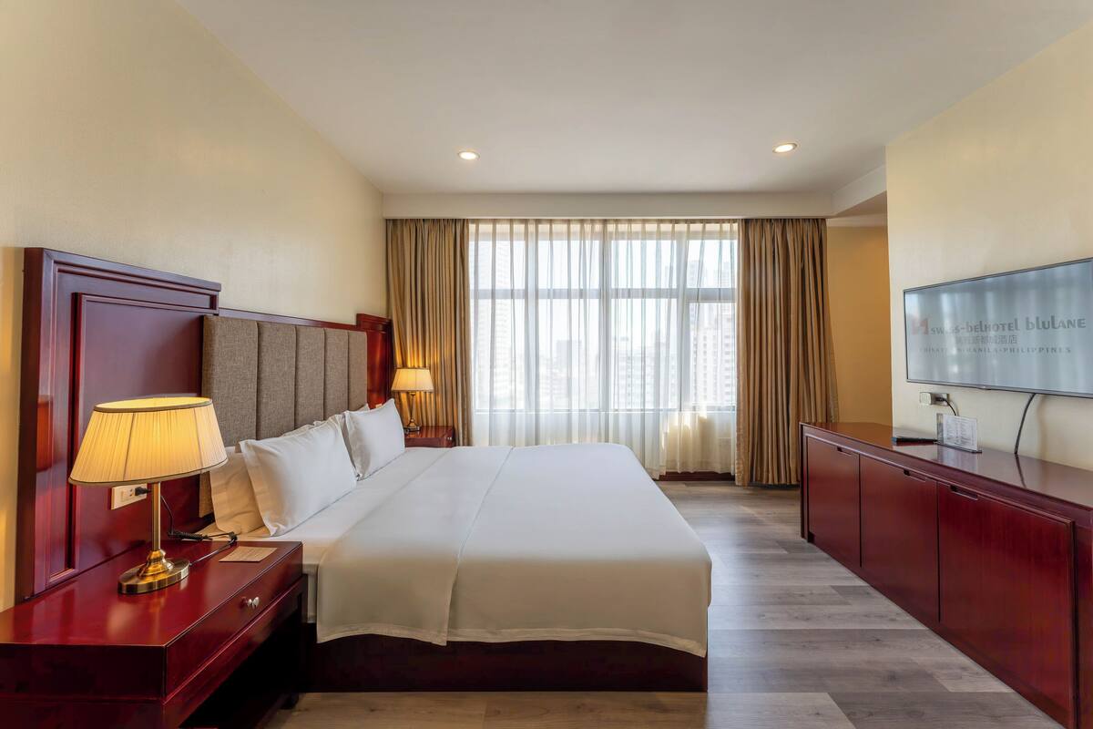 A hotel room with a large bed, a TV, a desk with a lamp, and a window with curtains.