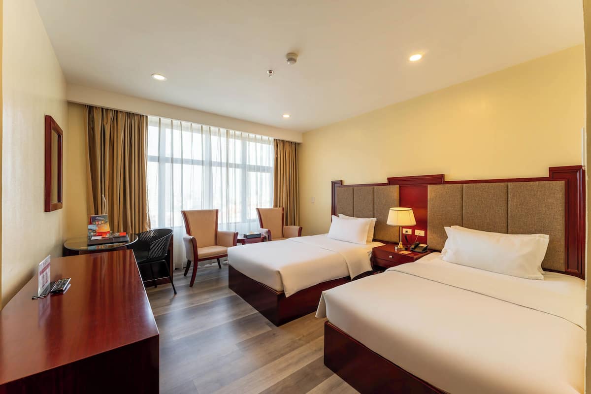 A hotel room with two beds, a desk, a chair, and a large window with curtains.