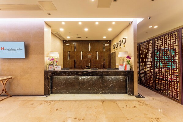 Zona de recepción de un hotel con un mostrador de mármol, dos lámparas de pared, un reloj y un panel decorativo de madera.