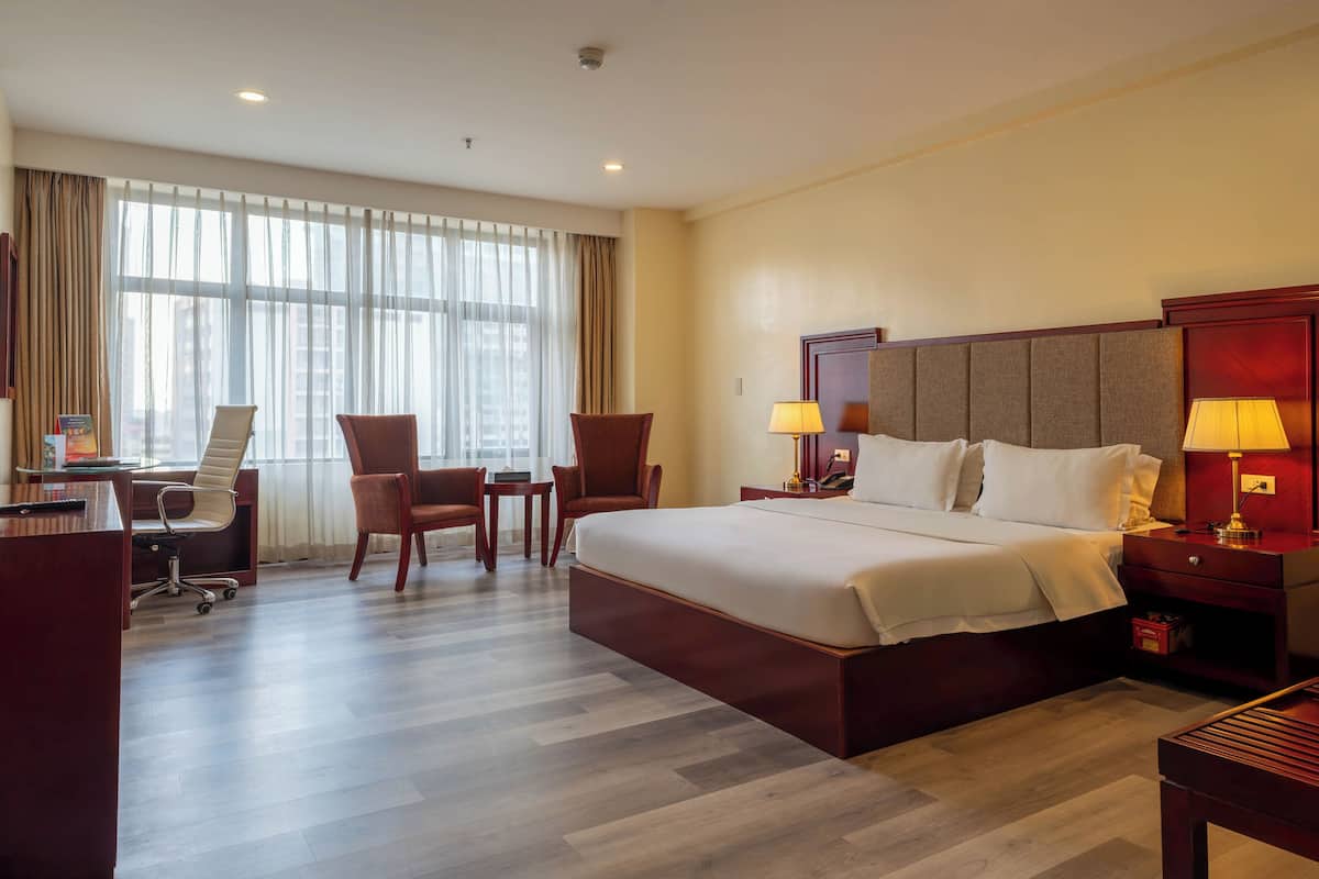 A hotel room with a large bed, two chairs, a desk, and a window with curtains.