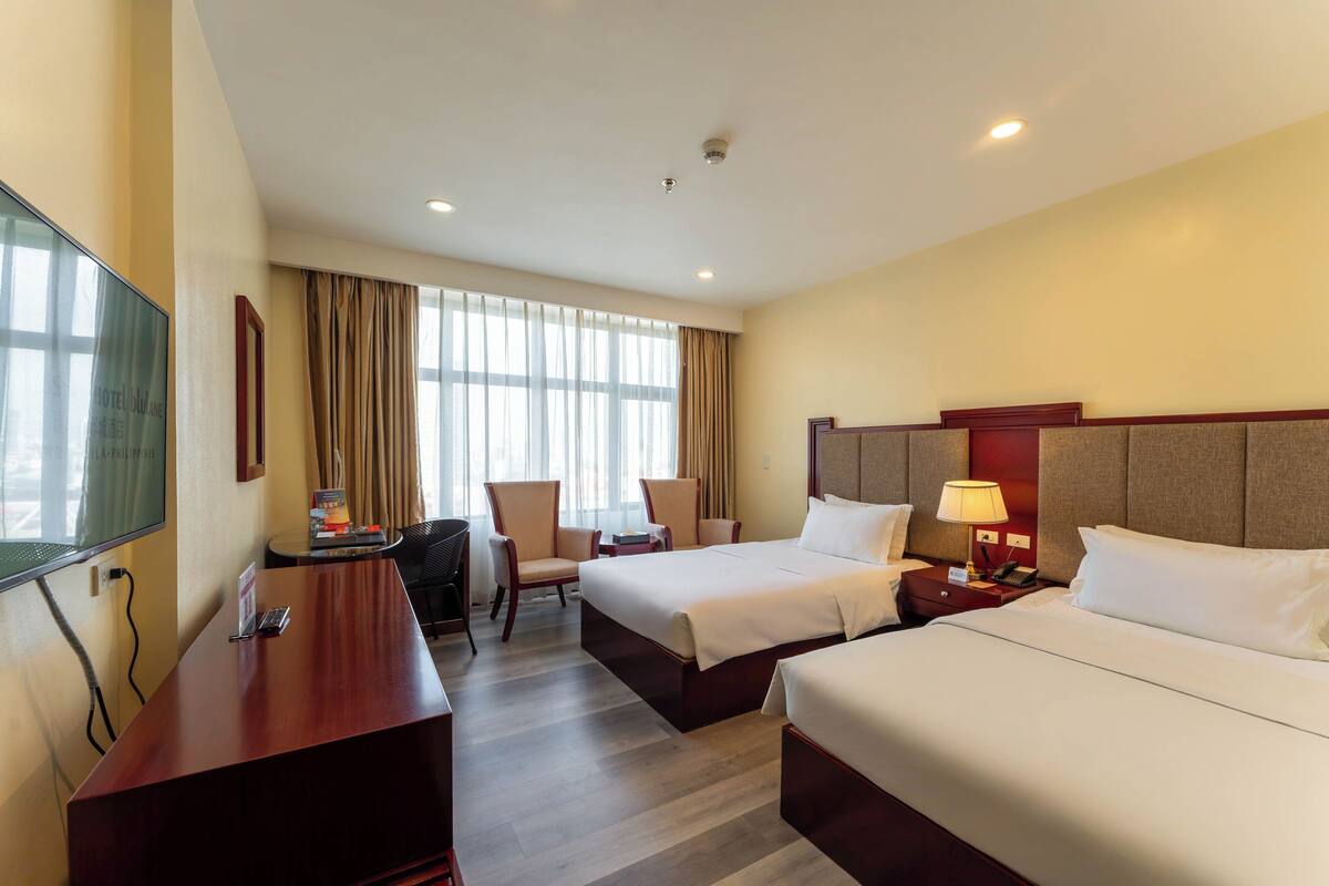 A hotel room with two beds, a desk, a chair, a TV, and a window with curtains.
