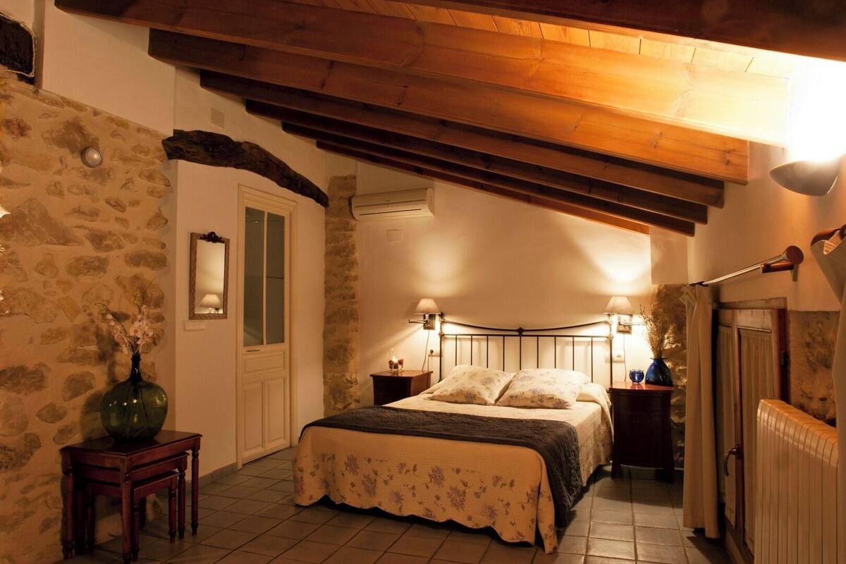A bedroom with a stone wall, wooden beams, a bed with floral bedding, bedside lamps, and a small table with a vase.