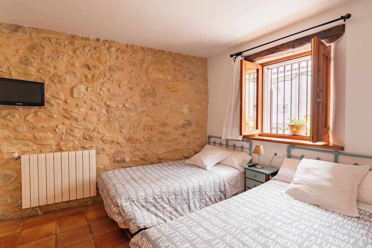 A bedroom with two beds, a stone wall, a window with a view, and a flat-screen TV mounted on the wall.