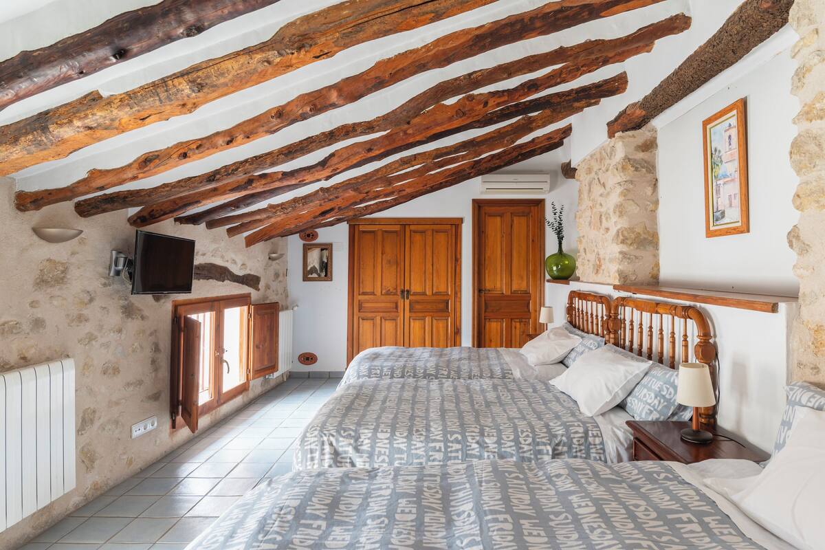 A bedroom with exposed wooden beams, a bed with patterned bedding, a bedside table with a lamp, and a television on the wall.