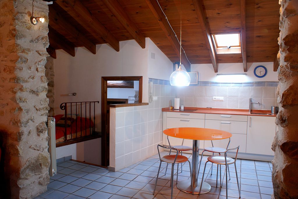 A rustic kitchen with a wooden ceiling, a round orange table with four chairs, and a stone wall.