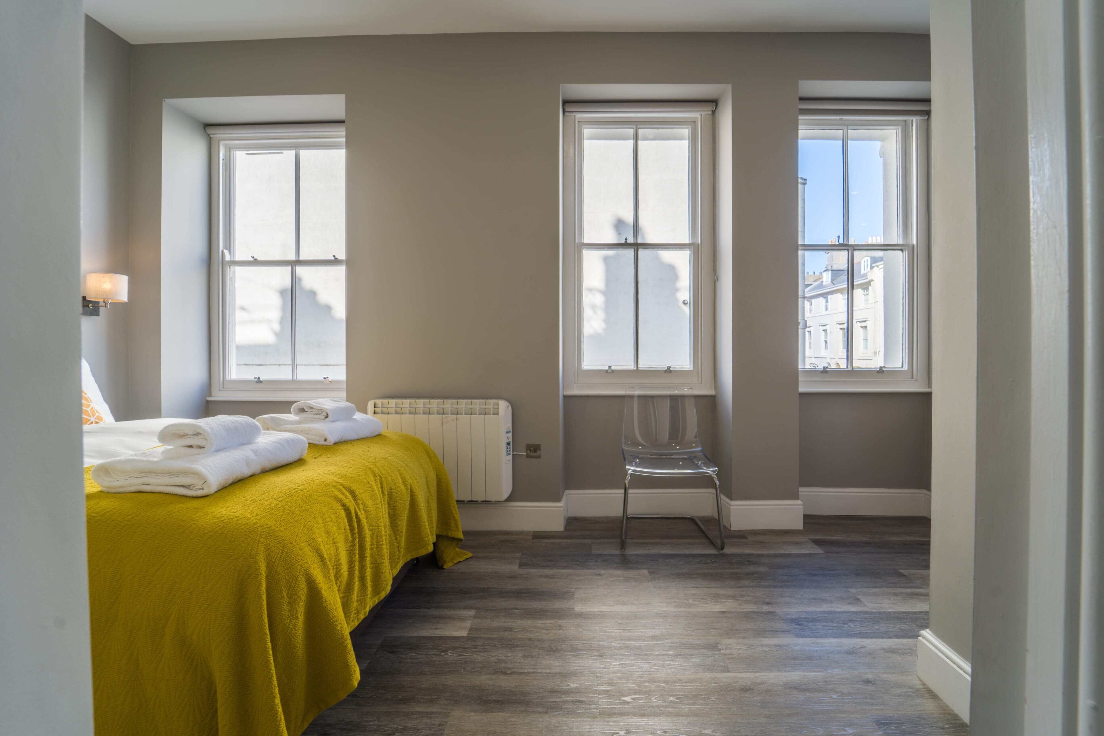 Un dormitorio con cama, dos ventanas, un radiador y una silla.