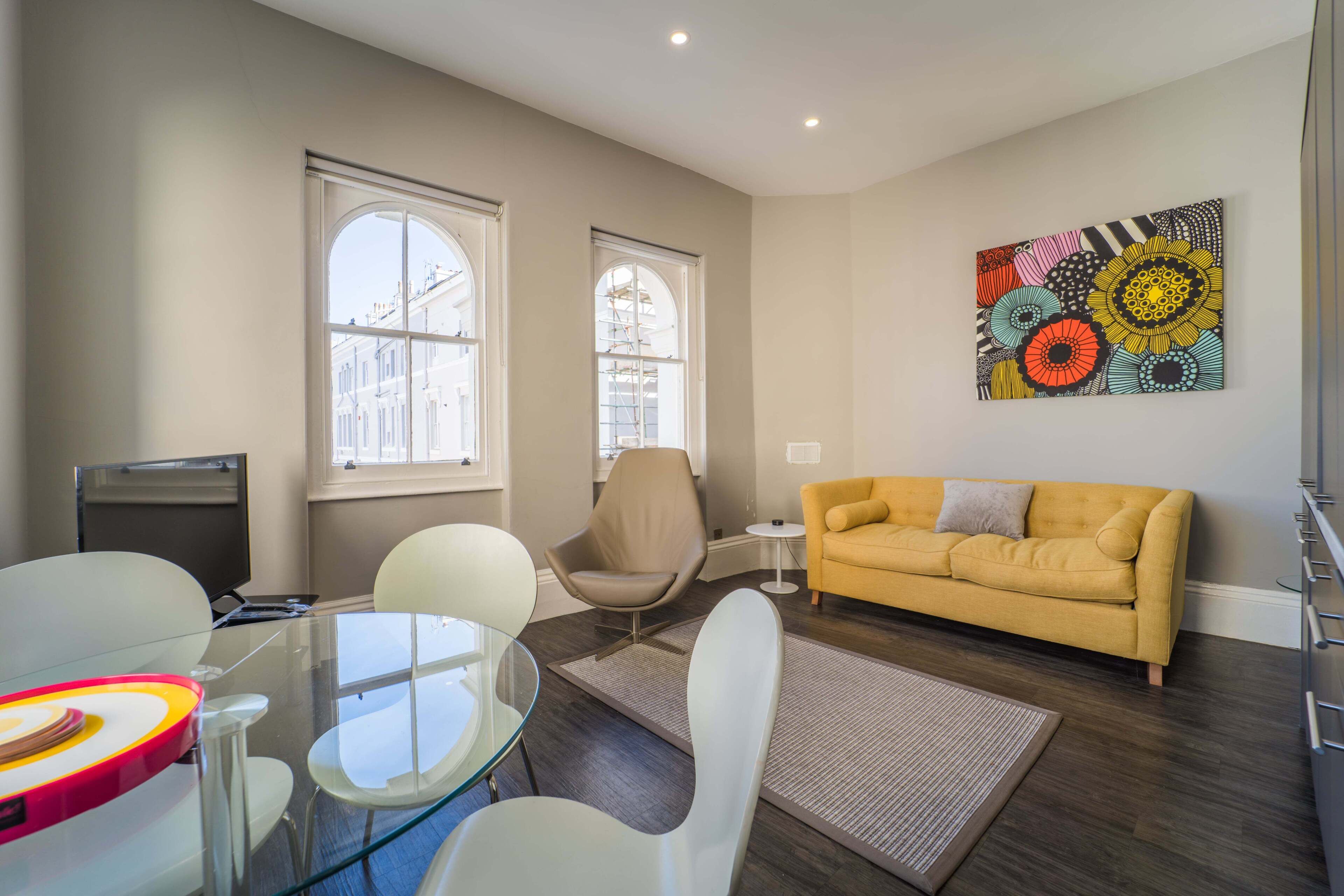 A modern living room with a glass table, white chairs, a yellow sofa, and a colorful abstract painting on the wall.