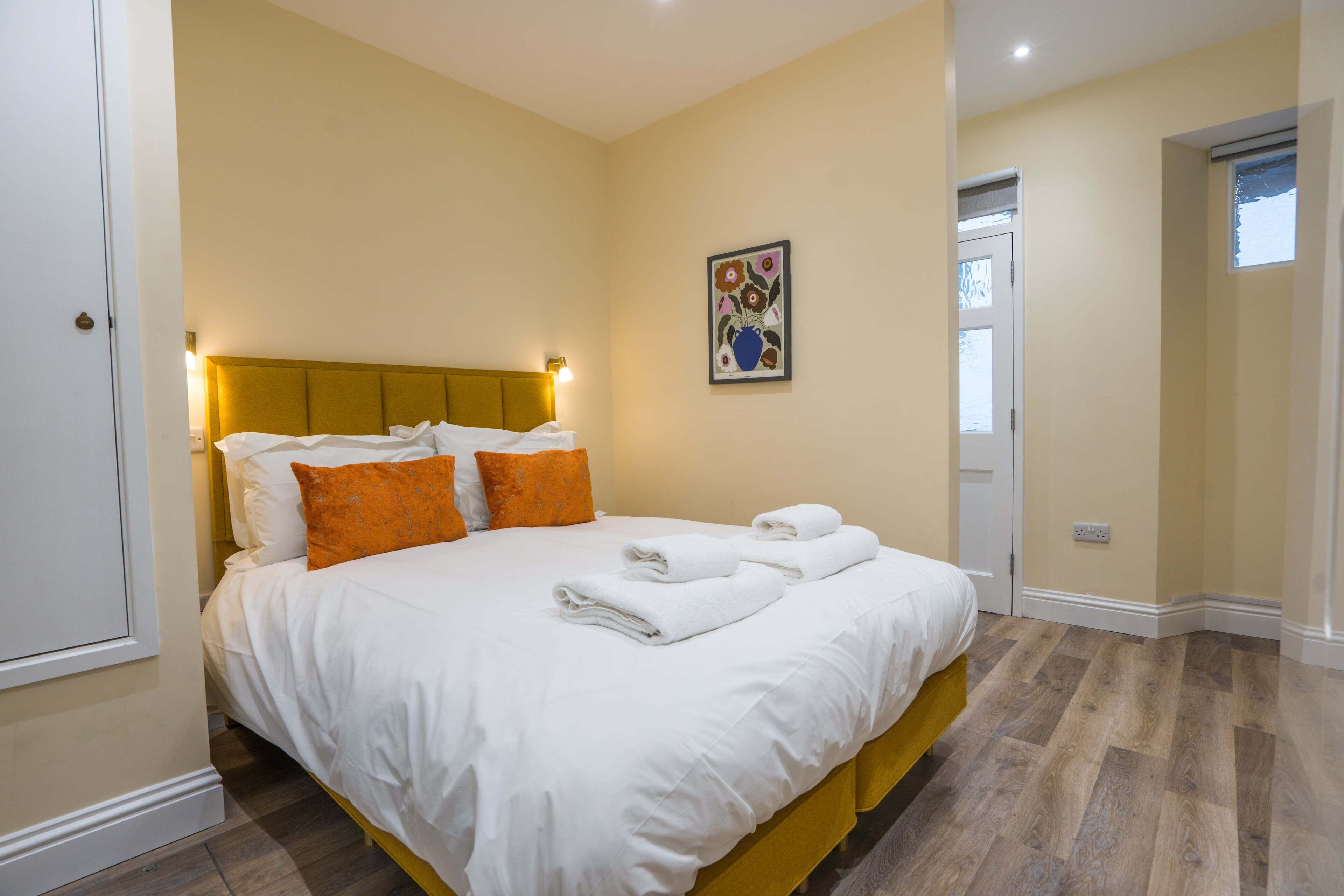 A neatly made bed with white and orange pillows, a headboard with integrated lighting, and a framed floral artwork on the wall.