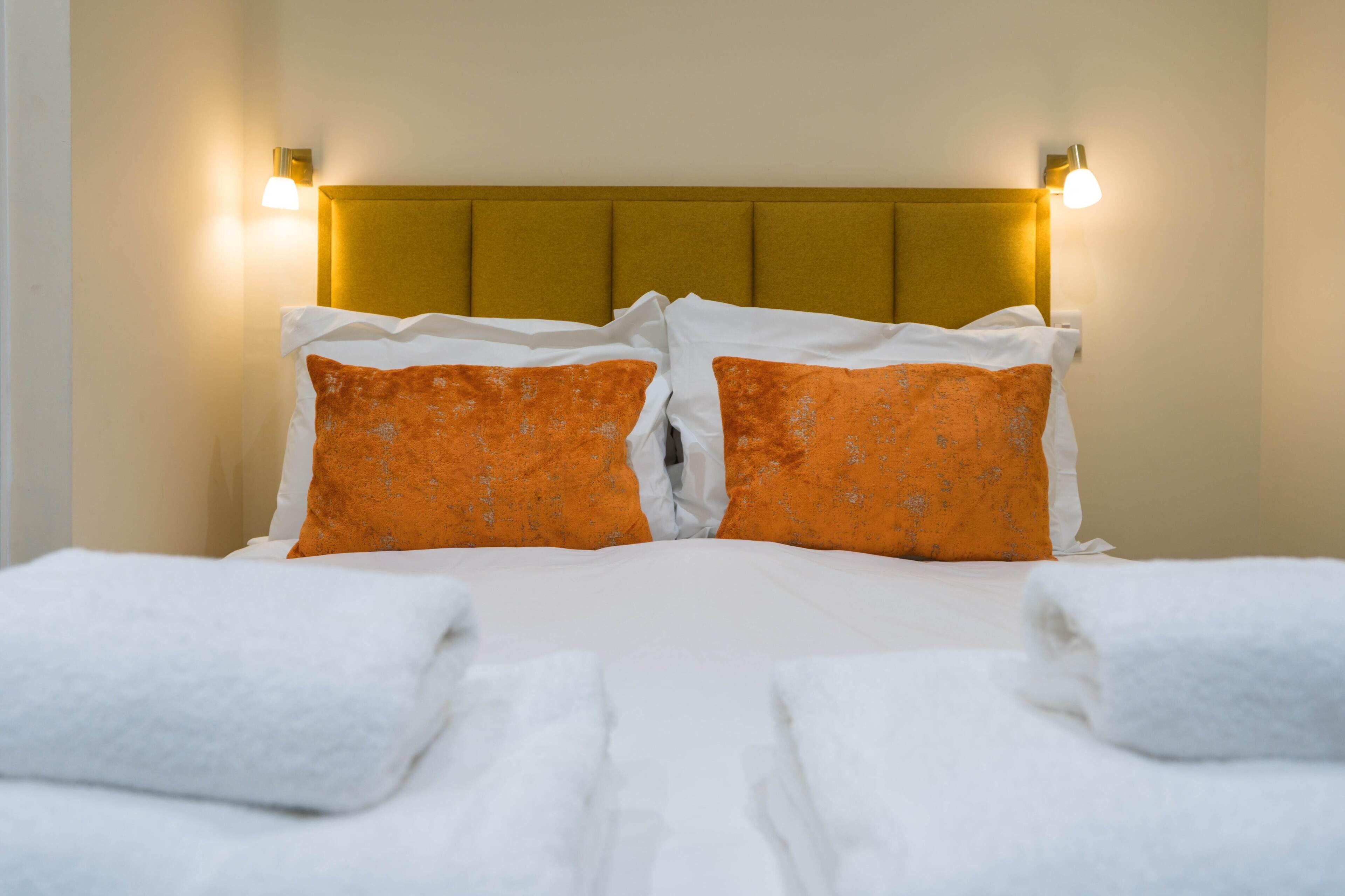 Un lit bien fait, agrémenté d’oreillers blancs et orange, d’une tête de lit dorée et de deux lampes fixées au mur.