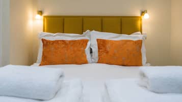 Un lit bien fait, agrémenté d’oreillers blancs et orange, d’une tête de lit dorée et de deux lampes fixées au mur.