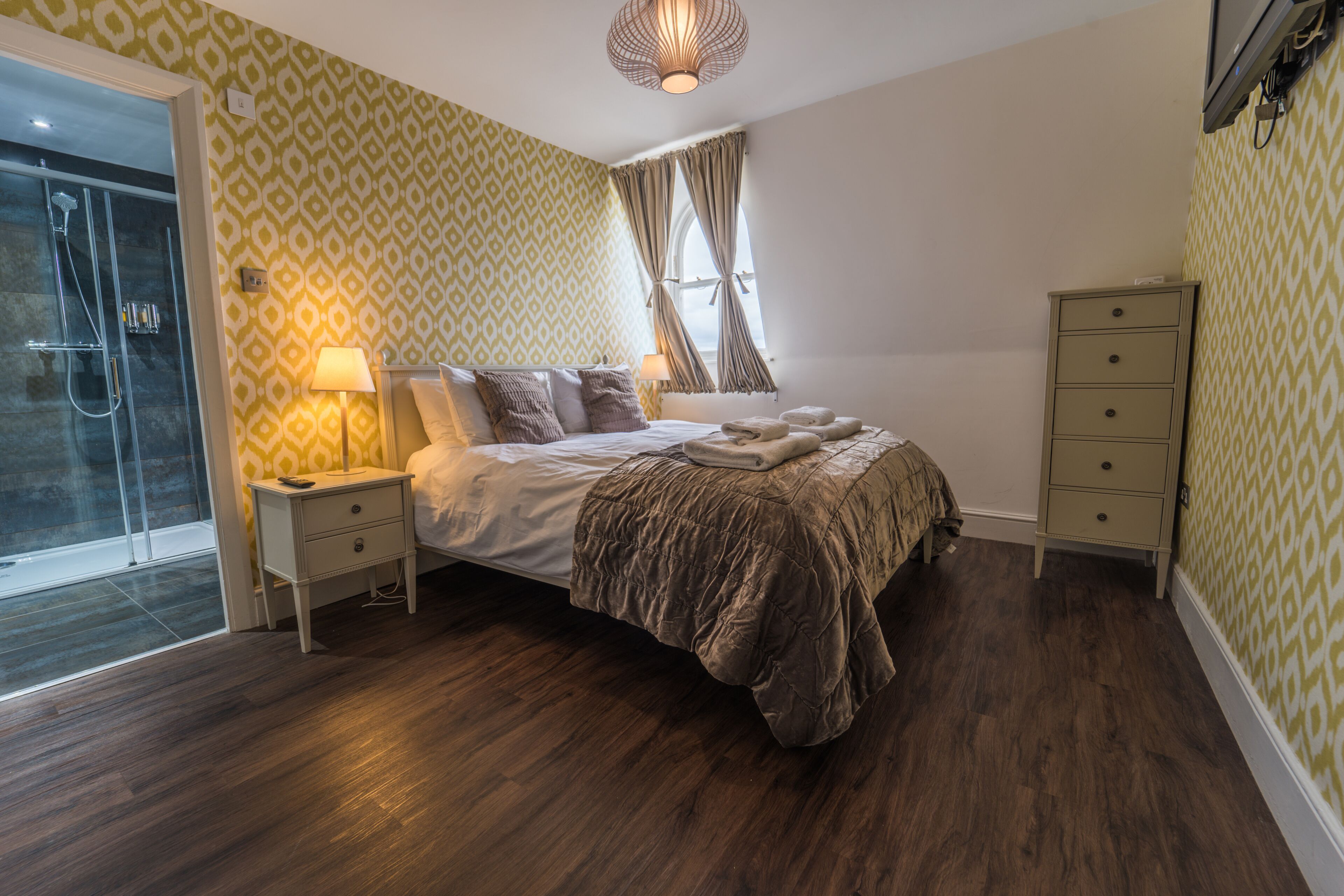 A bedroom with a bed, bedside tables, a wardrobe, a shower area, and patterned wallpaper.