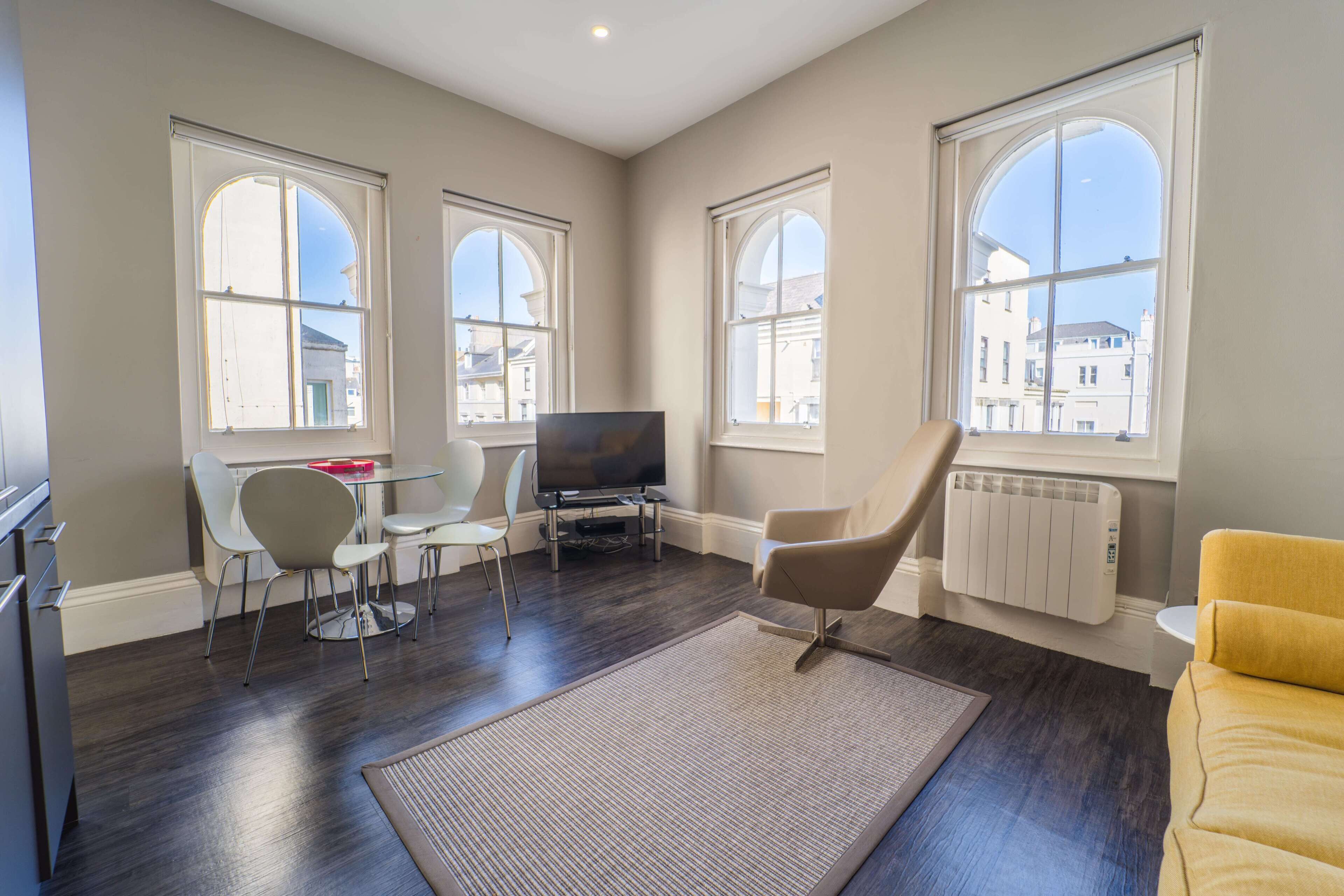 A modern living room with a flat-screen TV, a dining table with chairs, a sofa, and large windows with a city view.