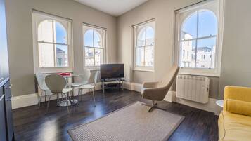 A modern living room with a flat-screen TV, a dining table with chairs, a sofa, and large windows with a city view.
