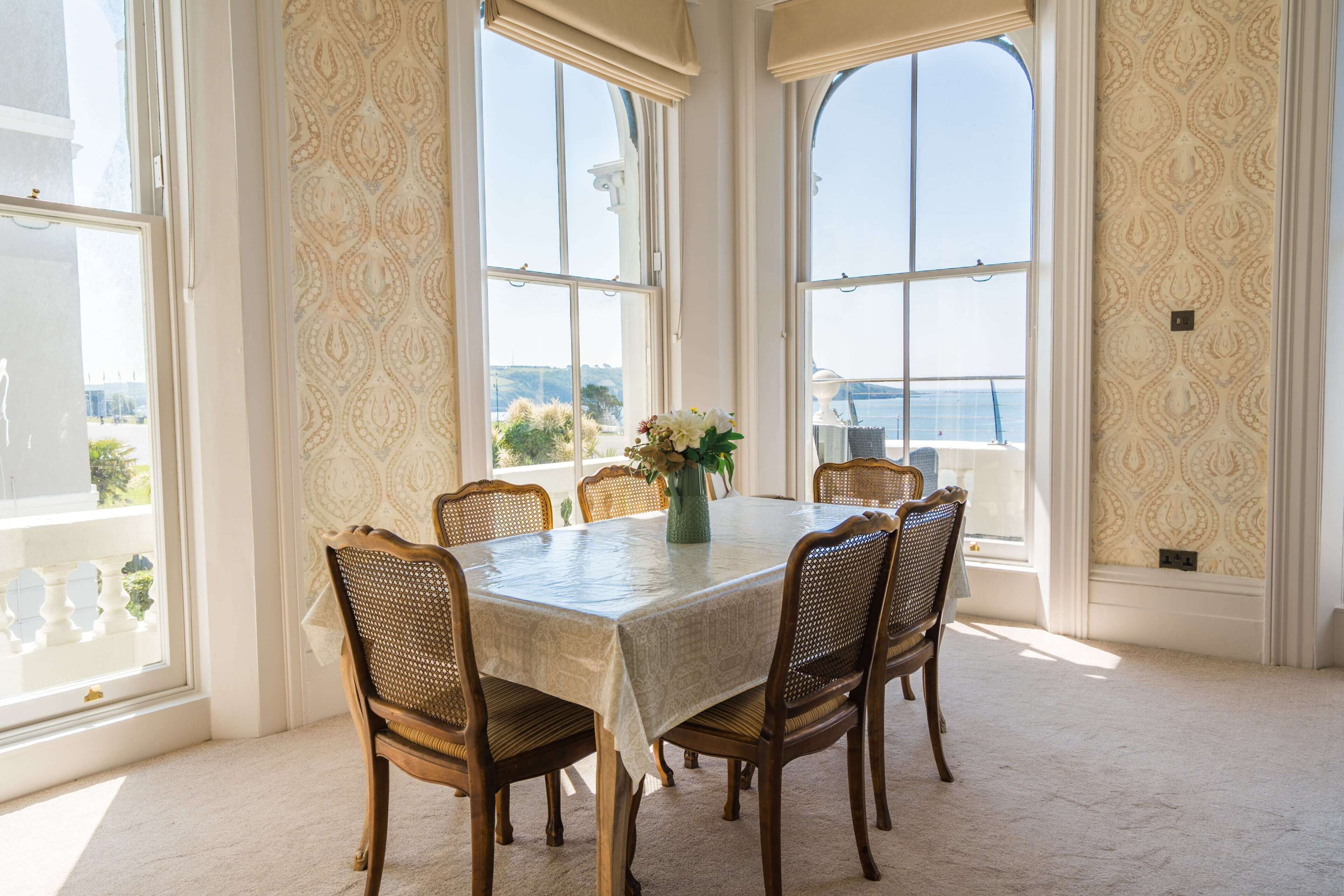 A dining area with a table set for four, surrounded by wicker chairs, large windows with a view, and patterned wallpaper.