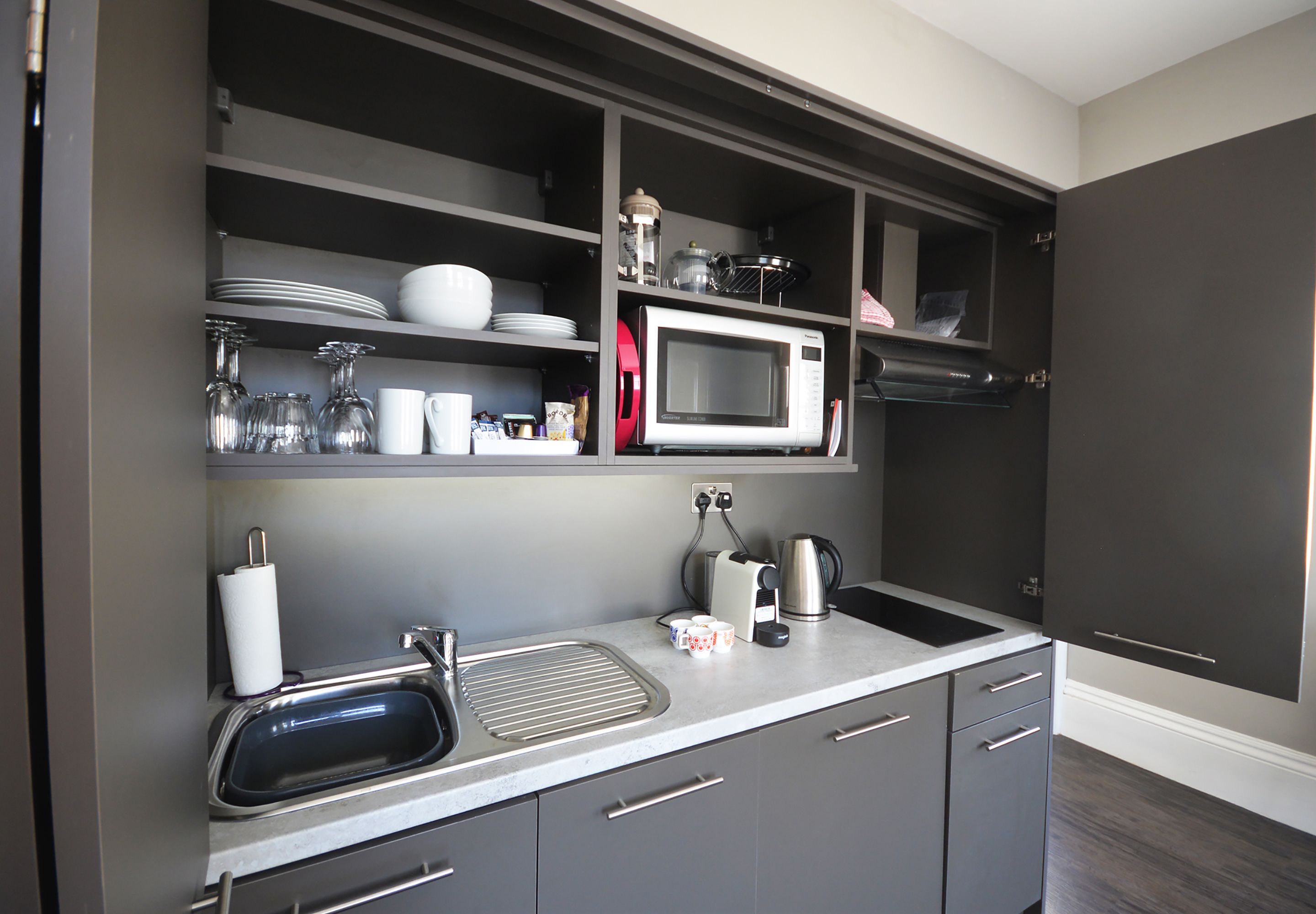 A modern kitchen with grey cabinets, a stainless steel sink, a microwave, and a kettle.
