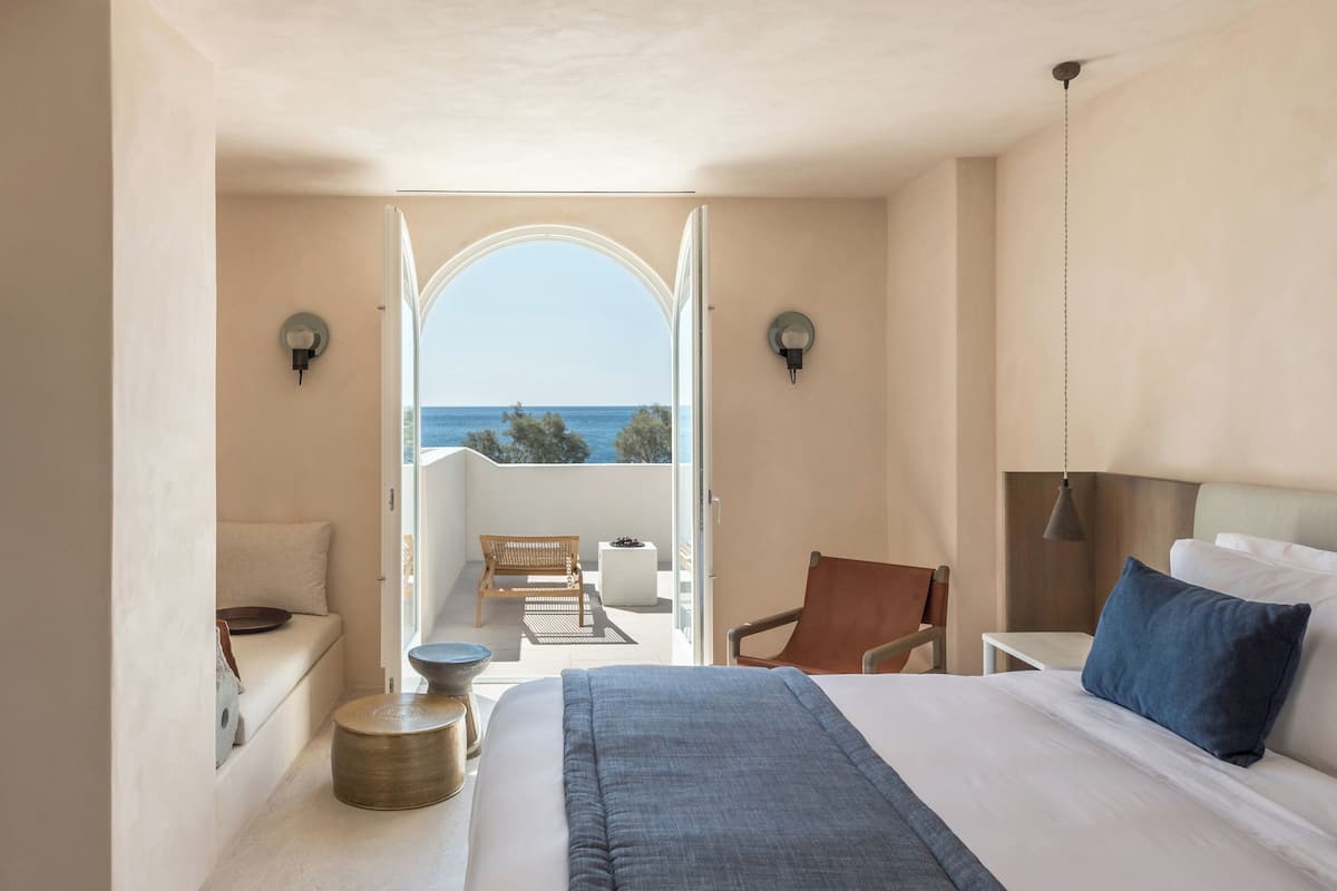 Un dormitorio con una cama grande, una silla, una mesita y vistas al mar a través de una puerta abierta.