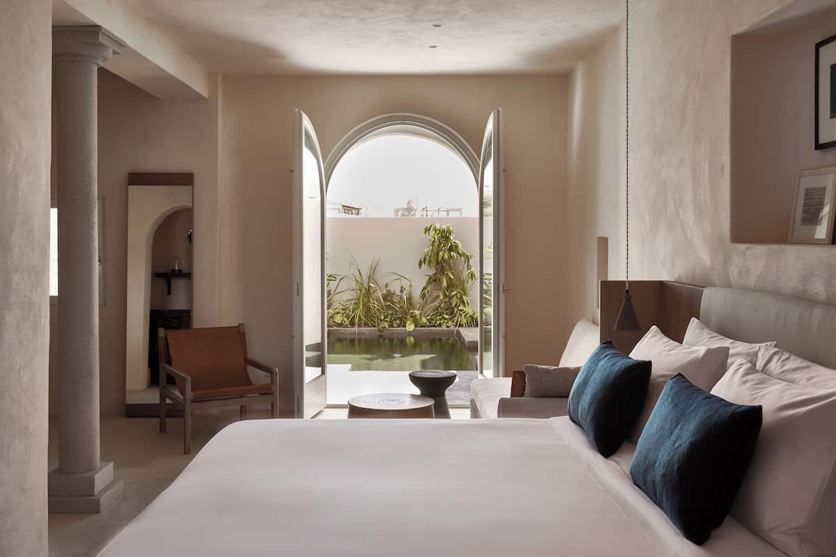 Habitación de hotel con una cama grande, vista a un jardín a través de un arco abierto y una mesita con un cuenco.