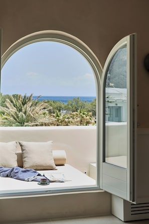 Ein Zimmer mit einem Bett, einem Fenster mit geöffneter Tür und Blick auf Meer und Grünfläche.