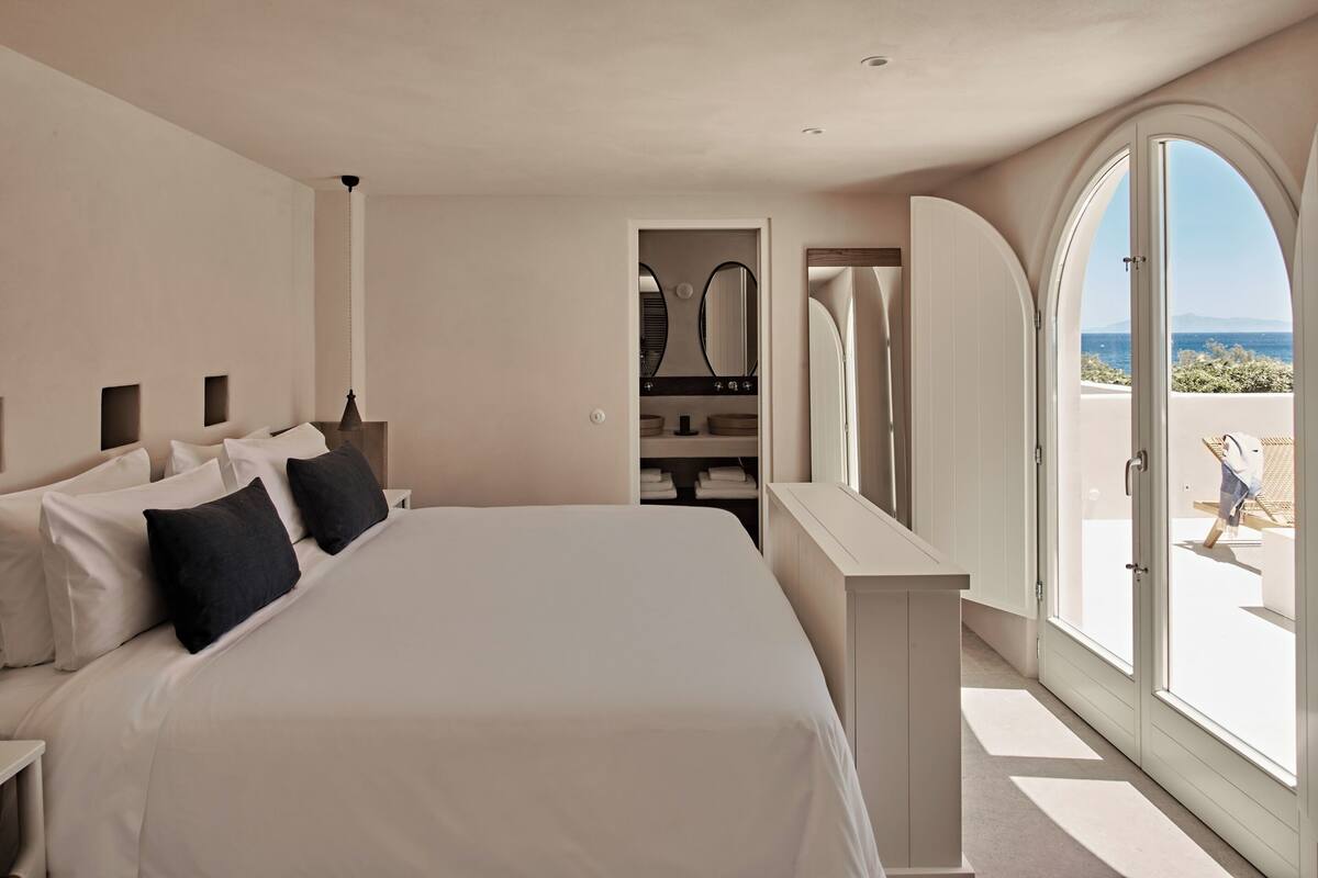 Un dormitorio con cama, mesita de noche, espejo y vista al mar a través de un amplio ventanal de arco.