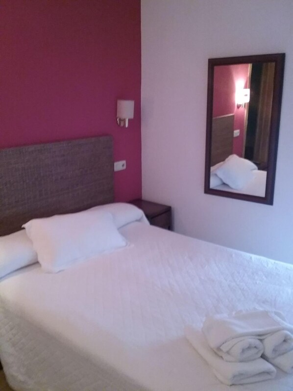 Una habitación de hotel con cama, mesita de noche, espejo montado en la pared y enchufe.