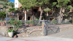 Un edificio de piedra con entrada protegida por una reja, rodeado de macetas con plantas y árboles.