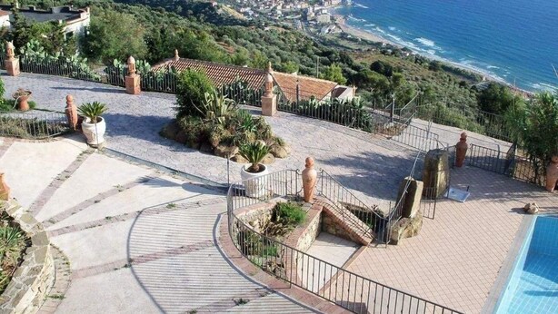 Un área de piscina en la azotea con vista al mar, rodeada de jardines escalonados y una valla.